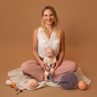 Carina Welzel sitzt im Schneidersitz auf einer Decke. Vor ihr liegen Arbeitsmaterialien für ihre Beratung: eine Babypuppe mit Plazenta- und Nabelschnurmodell, gehäkelte Modelle von Brüsten und Magengrößen zur Veranschaulichung von Stillmethoden und der kindlichen Magenentwicklung.
